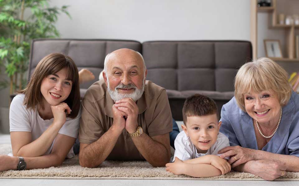 Deux couples de seniors allongés sur le sol, entourés de leurs petits-enfants, souriant et profitant de moments de complicité en famille.